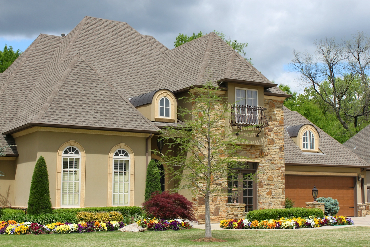 A two-story house in Spring, TX with a brown roof and beige exterior, featuring multiple gables and arched windows. The front yard includes a well-maintained garden with various colorful flowers and small bushes, and a young tree stands near the center. A double garage is on the right.