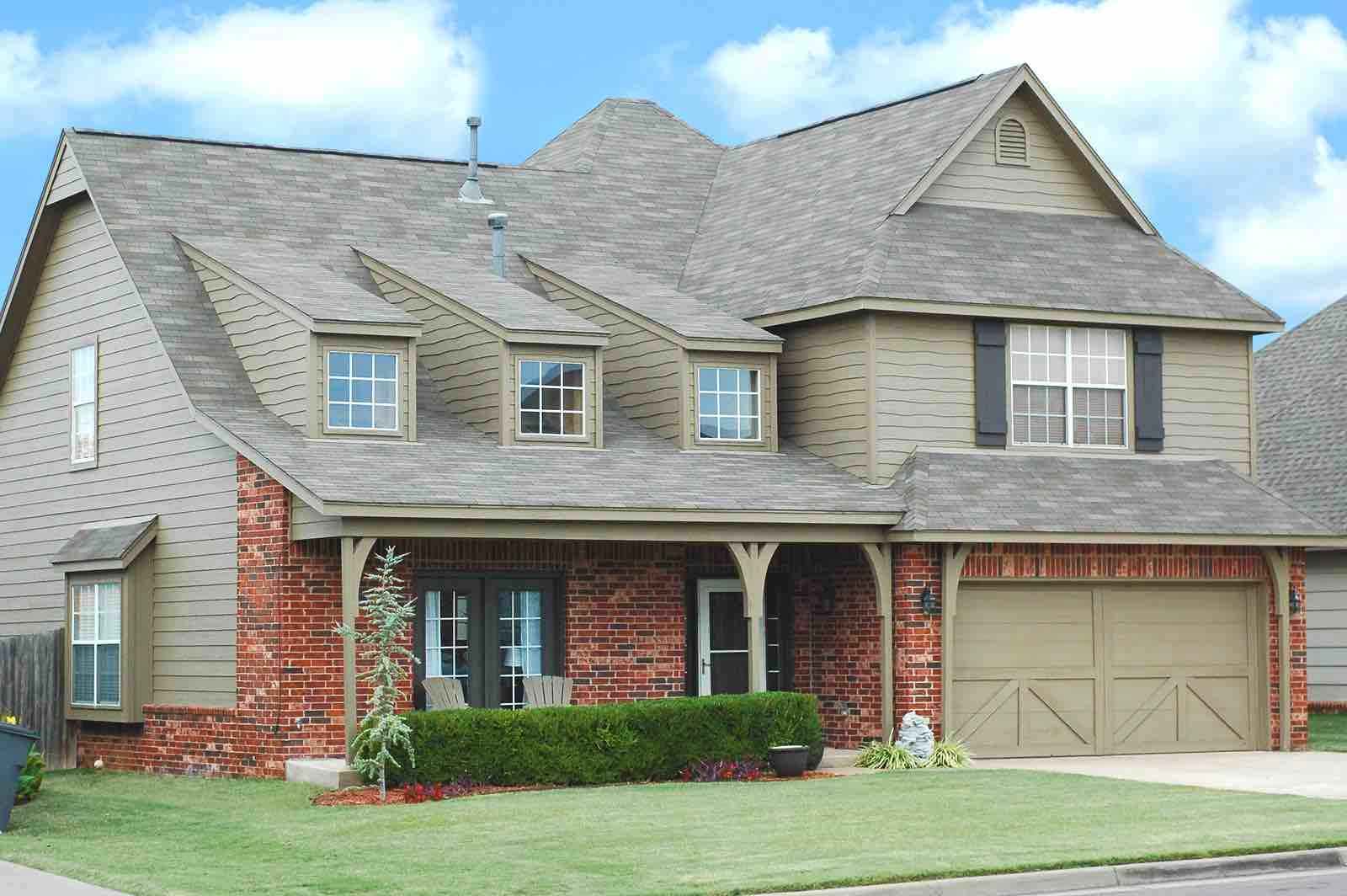 A two-story suburban house with a mix of red brick and beige siding. The house features a large garage, multiple windows, and a well-maintained front yard with green grass and shrubs. There is a covered porch with a few chairs in front of the entrance.