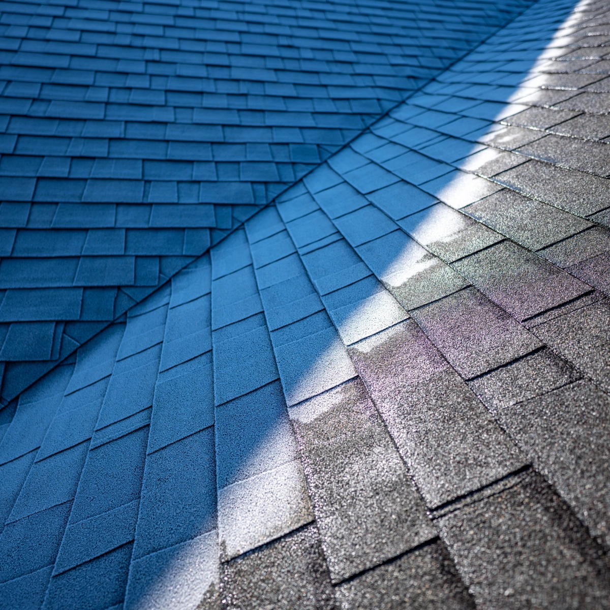 A close-up view of a roof with dark grey asphalt shingles. The roof, serviced recently by Pecan Grove roof repairs, has a valley where two sections meet, and sunlight creates a bright streak along the slope, casting a gradient of light and shadow across the shingles.