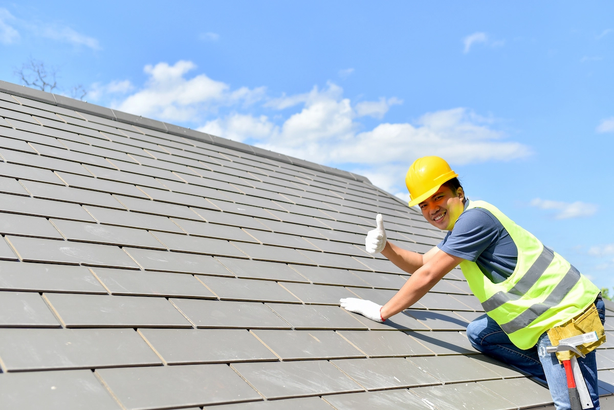 A construction worker wearing a yellow hard hat, a high-visibility vest, gloves, and jeans gives a thumbs up while kneeling on a roof covered with grey tiles. Beneath the clear blue sky with scattered clouds in the background, he showcases excellent Houston roofing workmanship.