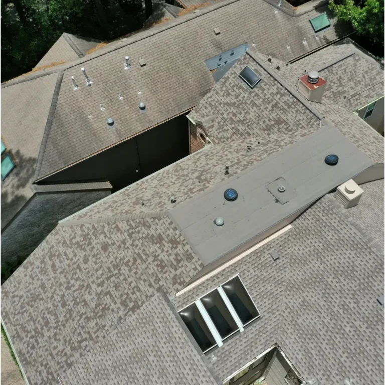 Aerial view of a large residential roof with multiple peaks, valleys, and sections. The roof is covered with light brown shingles and features various roof penetrations, including vents, skylights, and a chimney. Trees and greenery are visible around the house.