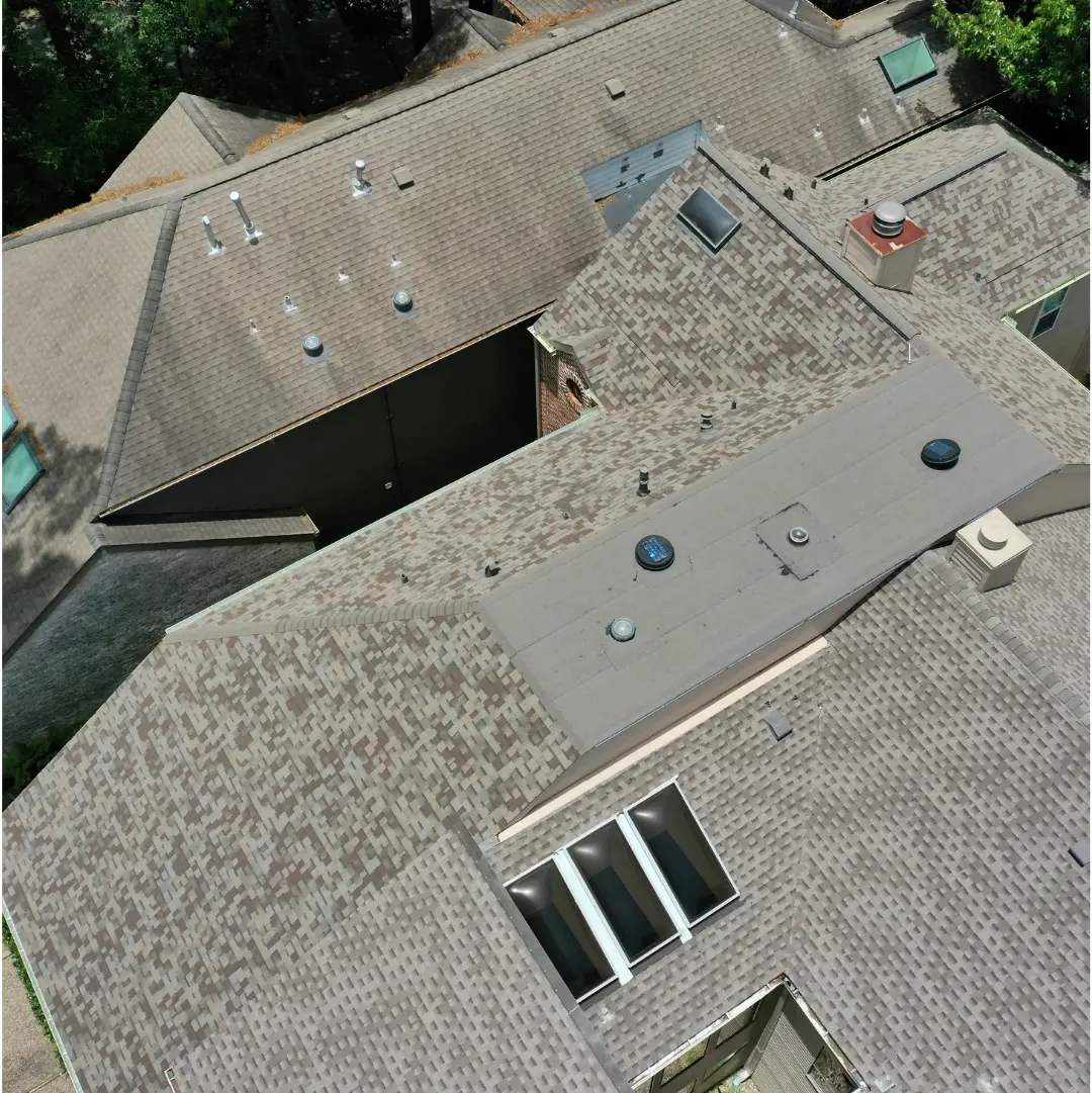 Aerial view of a large residential roof with multiple peaks, valleys, and sections. The roof is covered with light brown shingles and features various roof penetrations, including vents, skylights, and a chimney. Trees and greenery are visible around the house.
