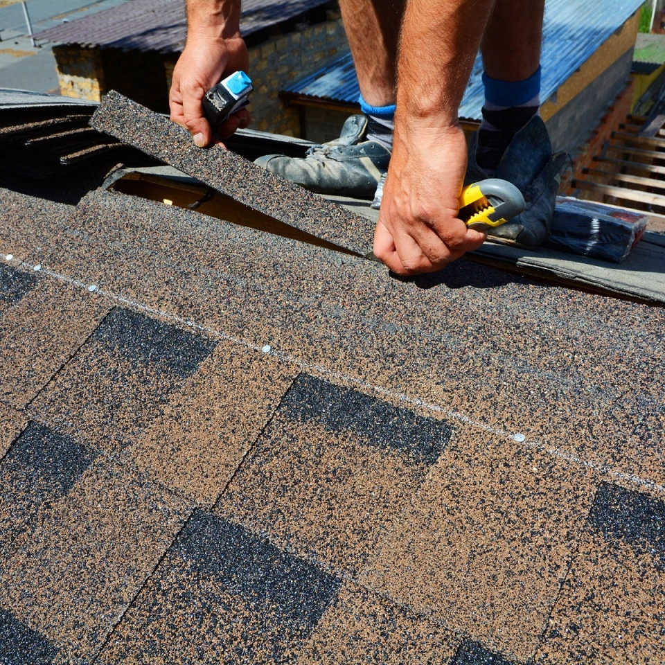 A Houston roofer's hands, with a tape measure and a utility knife, adjusting asphalt shingles on a pitched roof. The brown shingles have a speckled pattern, and part of a wooden structure can be seen in the background.