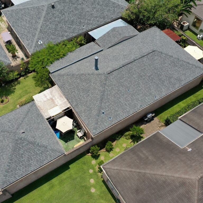 Aerial view of a residential neighborhood featuring houses with gray shingled roofs, neatly trimmed lawns, and some fenced patios. One patio includes an outdoor umbrella and seating area. Trees and shrubs are visible along the fences and beside houses, ensuring the charm of Houston's roof repair efforts.