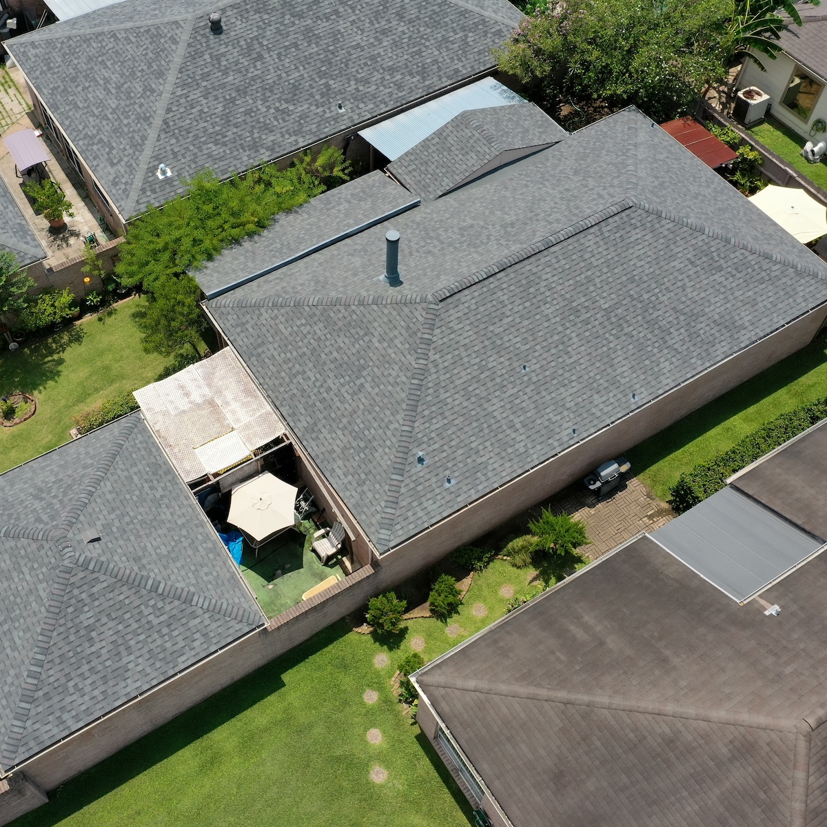 Aerial view of a residential neighborhood featuring houses with gray shingled roofs, neatly trimmed lawns, and some fenced patios. One patio includes an outdoor umbrella and seating area. Trees and shrubs are visible along the fences and beside houses, ensuring the charm of Houston's roof repair efforts.