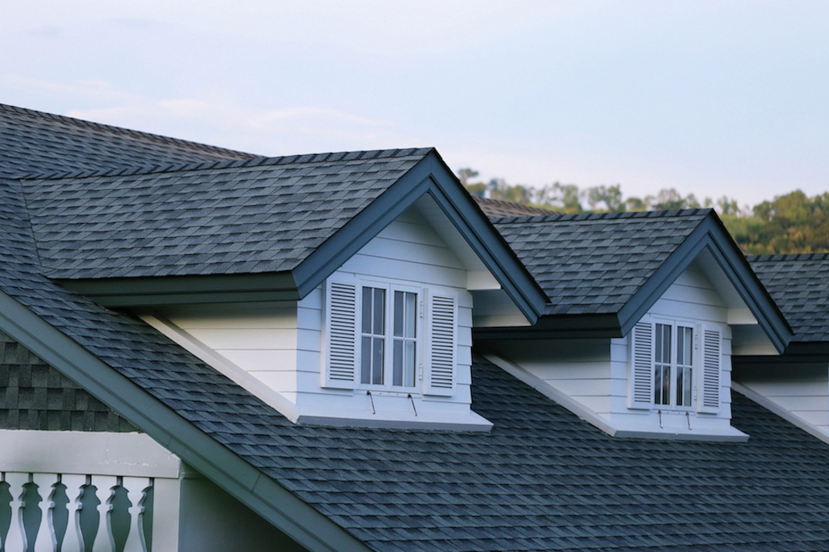 A close-up view of a house roof featuring two white dormer windows with shutters, set against a backdrop of a clear blue sky and tree line. The roof, showcasing expert Houston roofing craftsmanship, is covered with grey shingles, and there is a white railing visible on the left side of the image.
