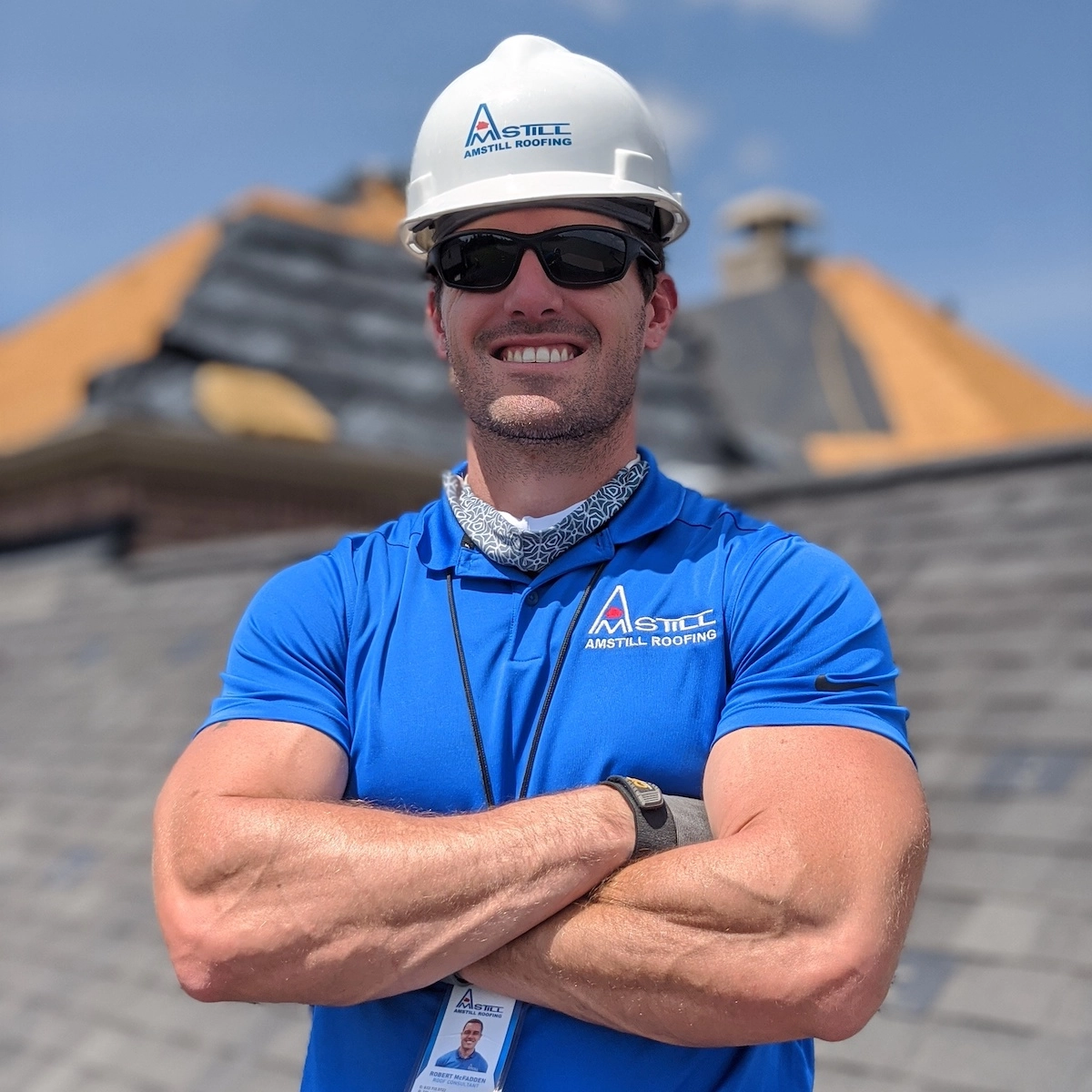 A smiling man wearing a white hard hat and blue polo shirt with the company logo "Amstill Roofing" stands outdoors in front of a building with a slate roof. He is crossing his arms and wearing black sunglasses and a lanyard, showcasing their expert four corners roof replacement service under a clear blue sky.