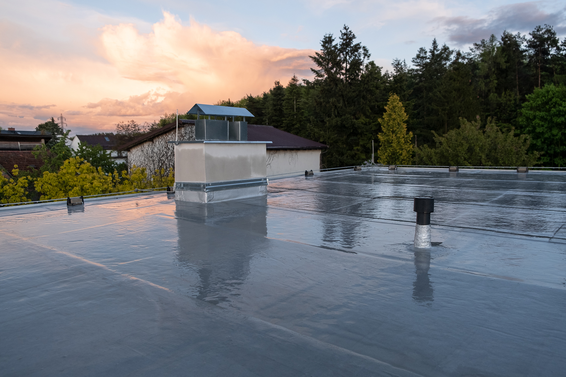 A flat rooftop covered with a smooth, reflective surface is shown. The roof has a vent stack, a chimney with metal flashing, and several other fixtures. Trees and a house are visible in the background under a partly cloudy sky during sunset.