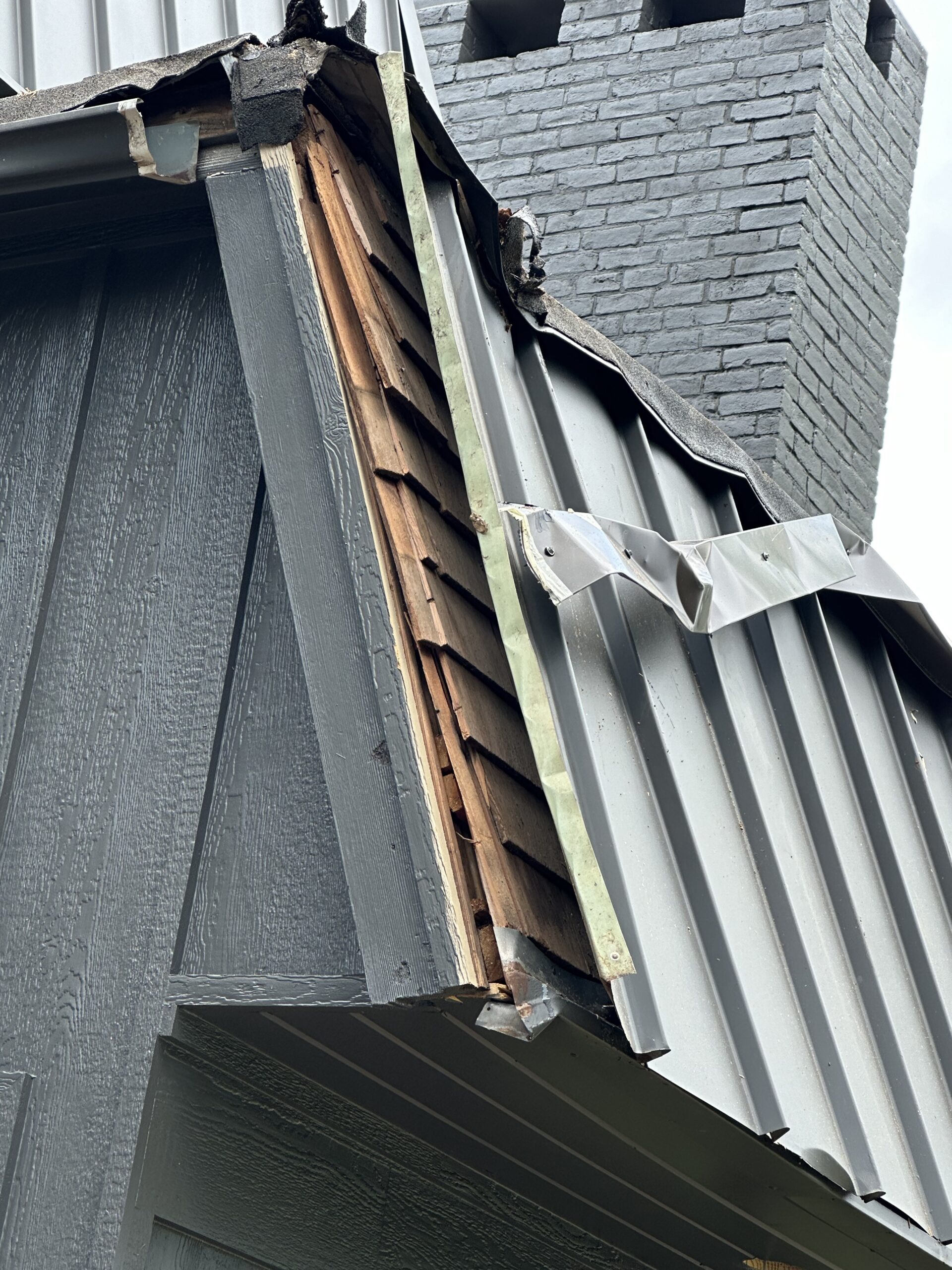 A close-up view of a damaged area on the side of a house, revealing exposed wood beneath the torn metal siding near a chimney. The metal siding is peeled back, and the underlying structure appears compromised. The chimney is constructed of gray bricks.