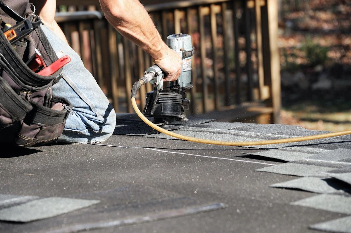 A person is installing shingles on a roof using a nail gun as part of a larger pecan grove roof replacement. The individual, equipped with a tool belt full of various tools, works diligently while a wooden railing stands visible in the background.