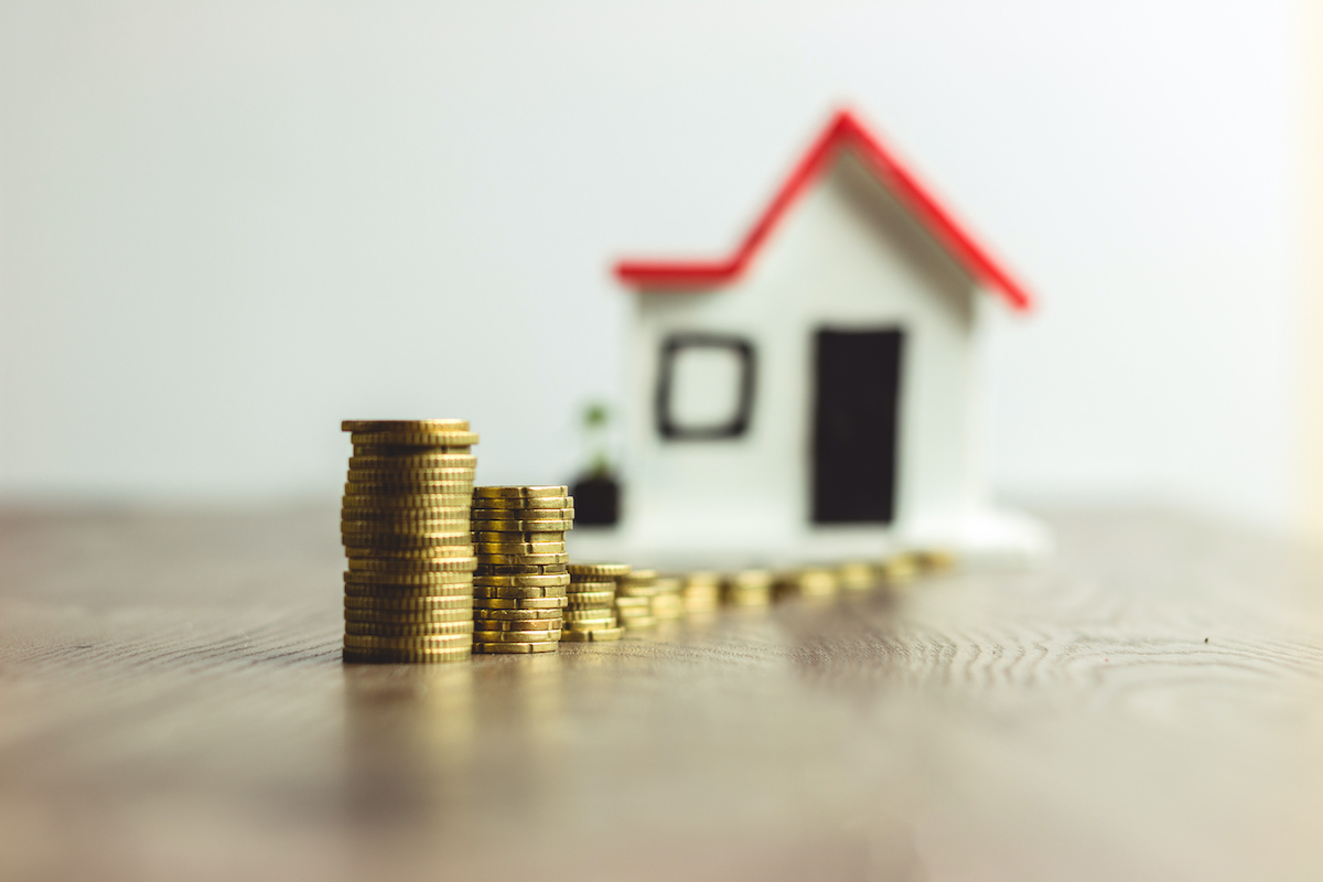 A row of stacked gold coins leads to a small, blurred model of a house with a red roof and white walls—a subtle reminder of the value in investing in quality services like Houston roof replacement. The coins are arranged in ascending height, creating a visual path toward the house against a softly focused background.