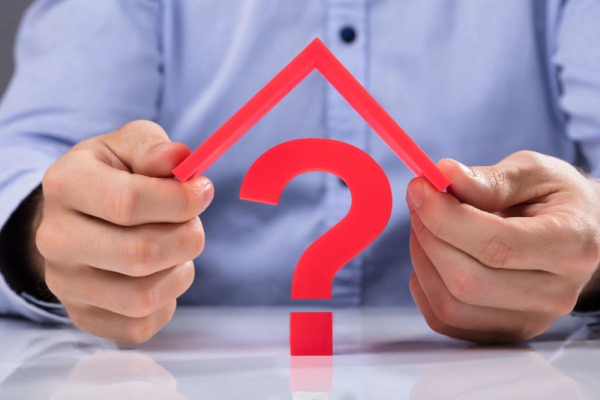 A person wearing a light blue shirt holds up a red house-shaped frame with a question mark inside it, suggesting uncertainty or inquiry about housing or real estate matters, possibly even Richmond roofing. Only the person's hands and part of the shirt are visible.