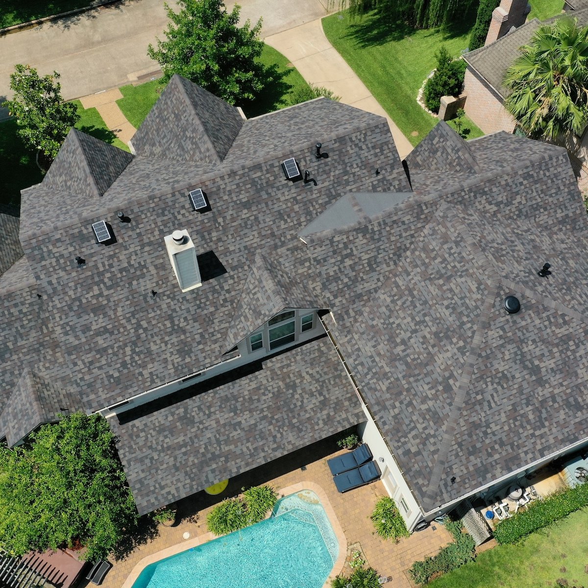 Aerial view of a residential property featuring a house with a dark gray, pecan grove roof replacement and a backyard with a swimming pool. The yard has neatly maintained greenery surrounding the pool, and the house has multiple roof angles and skylights.