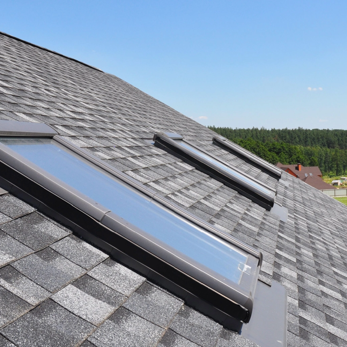 A Richmond roof replacement showcases a sloped roof with grey shingles and three skylights aligned in a row. The skylights are rectangular with dark frames, set against a clear blue sky and part of a residential neighborhood with green trees.