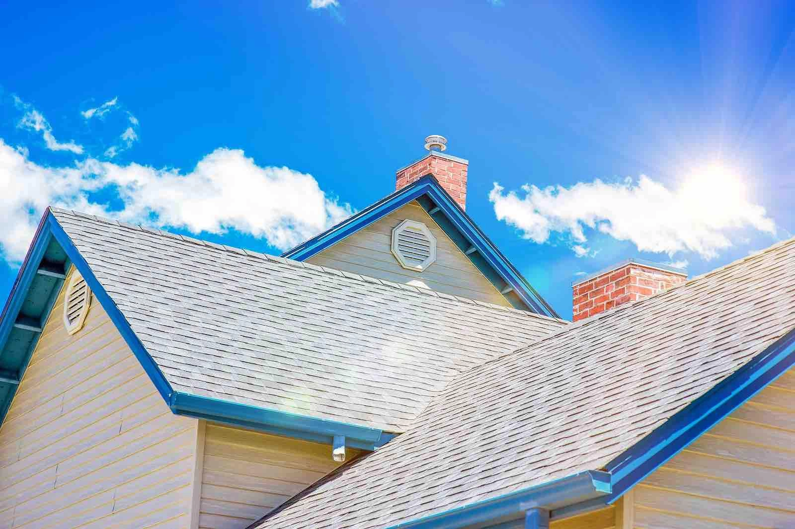 A close-up of a house roof with light blue trim under a bright, sunny sky. The roof is made of gray shingles, and there are two brick chimneys visible. The sun is shining brightly, with light clouds scattered in the blue sky.