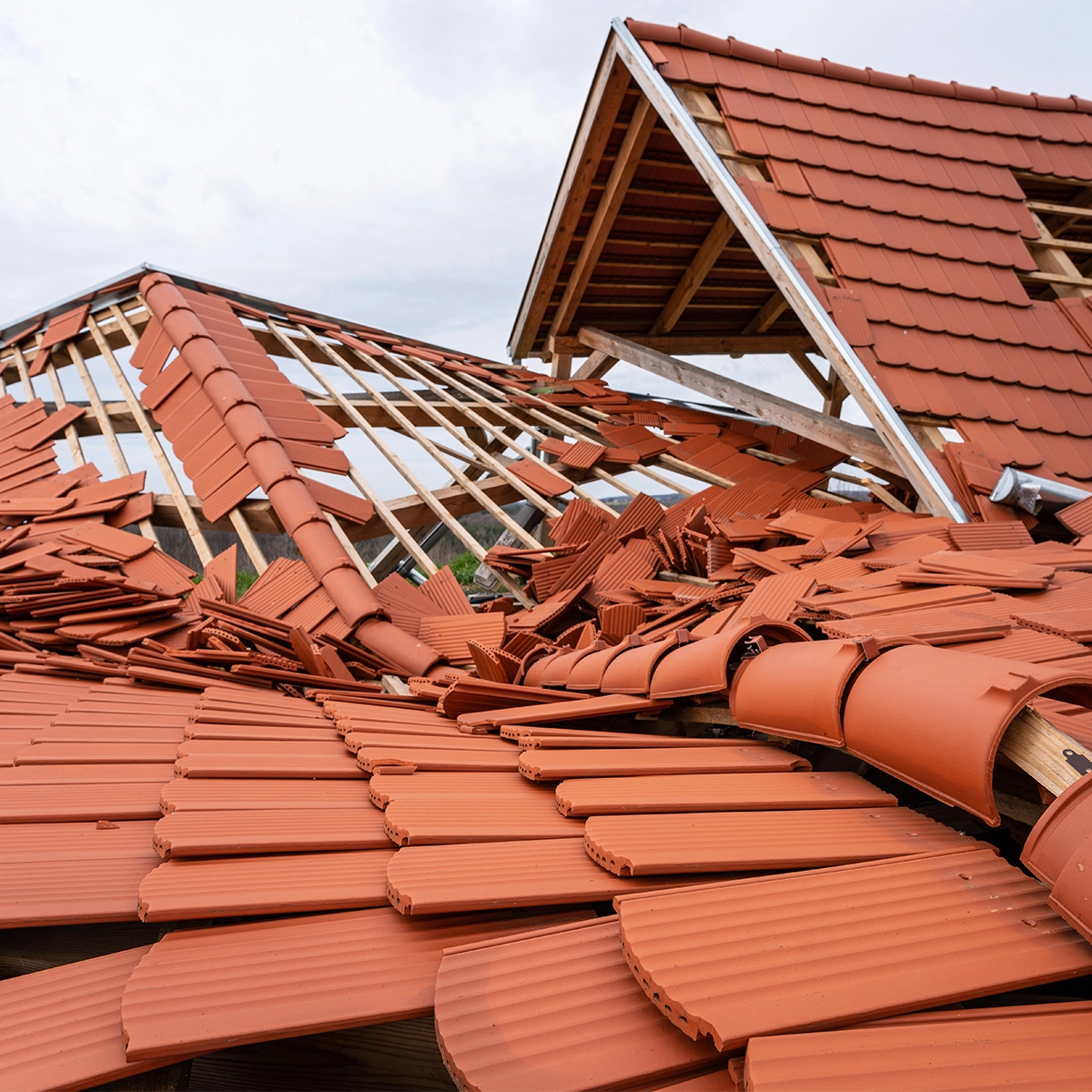 A collapsed red tile roof with wooden structural beams exposed in Sugar Land. The tiles are scattered and broken, parts of the roof have caved in, revealing extensive damage. The overcast sky enhances the scene's somber mood, clearly indicating the urgent need for a roof replacement.
