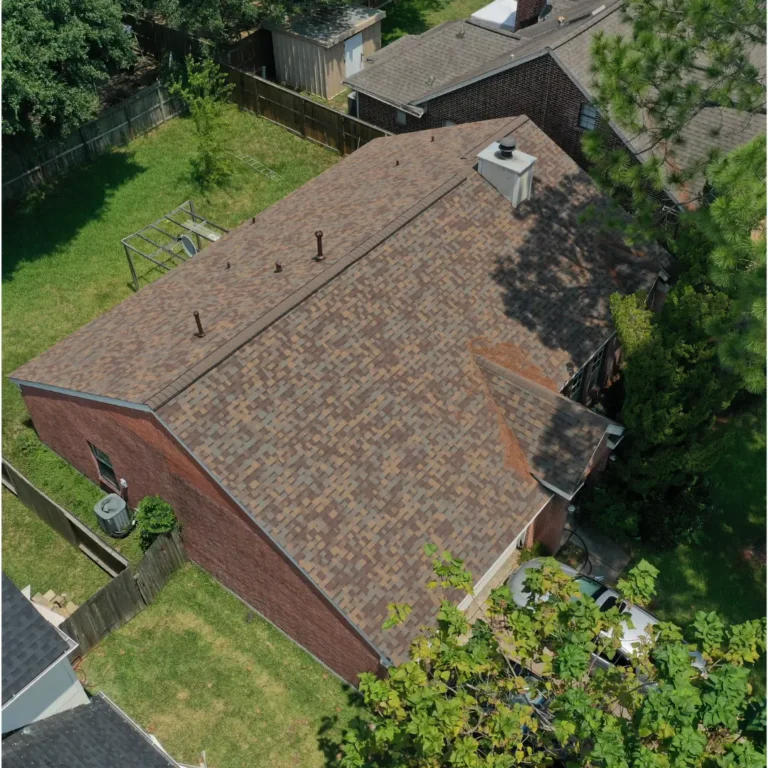 An aerial view of a suburban house with a brown shingle roof and red brick exterior. The backyard is fenced and includes a small patio and green lawn. Neighboring houses with similar architecture are also visible. Trees and greenery surround the area.