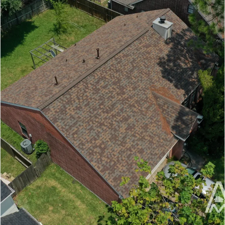 Aerial view of a residential house with a multi-brown shingle roof in good condition, surrounded by a fenced yard with green grass and trees. The house appears to be in a suburban neighborhood on a sunny day.