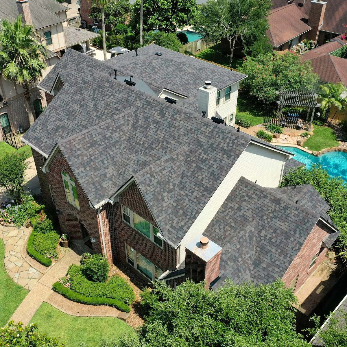 Aerial view of a suburban neighborhood featuring a large brick house with a dark shingle roof, recently upgraded through Richmond Roof Replacement. The house is surrounded by well-maintained landscaping, including a green lawn, bushes, and a stone pathway. A blue swimming pool and surrounding greenery are visible in the background.