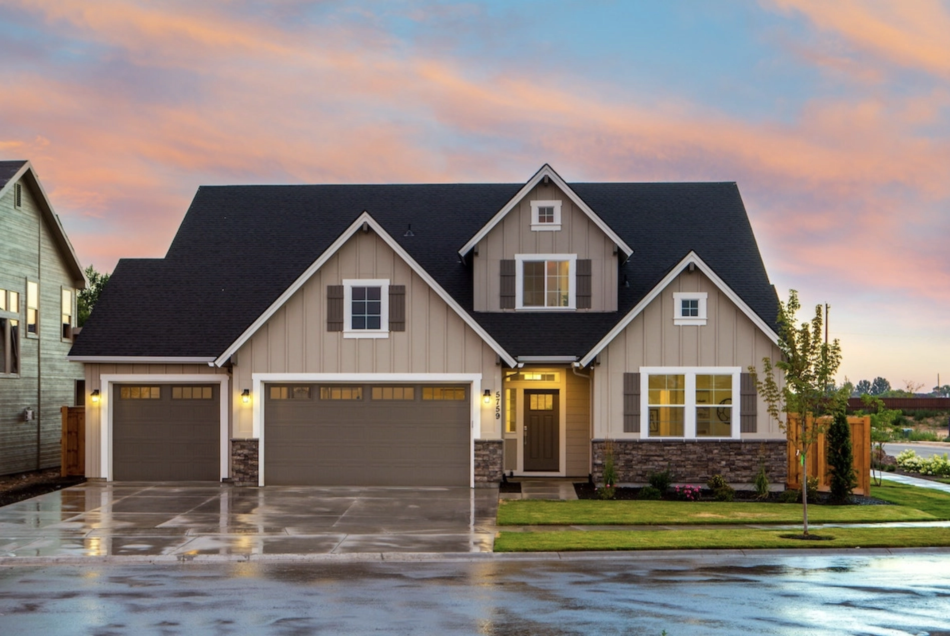 A modern suburban house at sunset, featuring a grey and white color scheme. The home has three garage doors, windows with shutters, and a covered front porch. The yard is well-kept with green grass and a small tree. The driveway and street are wet from rain.