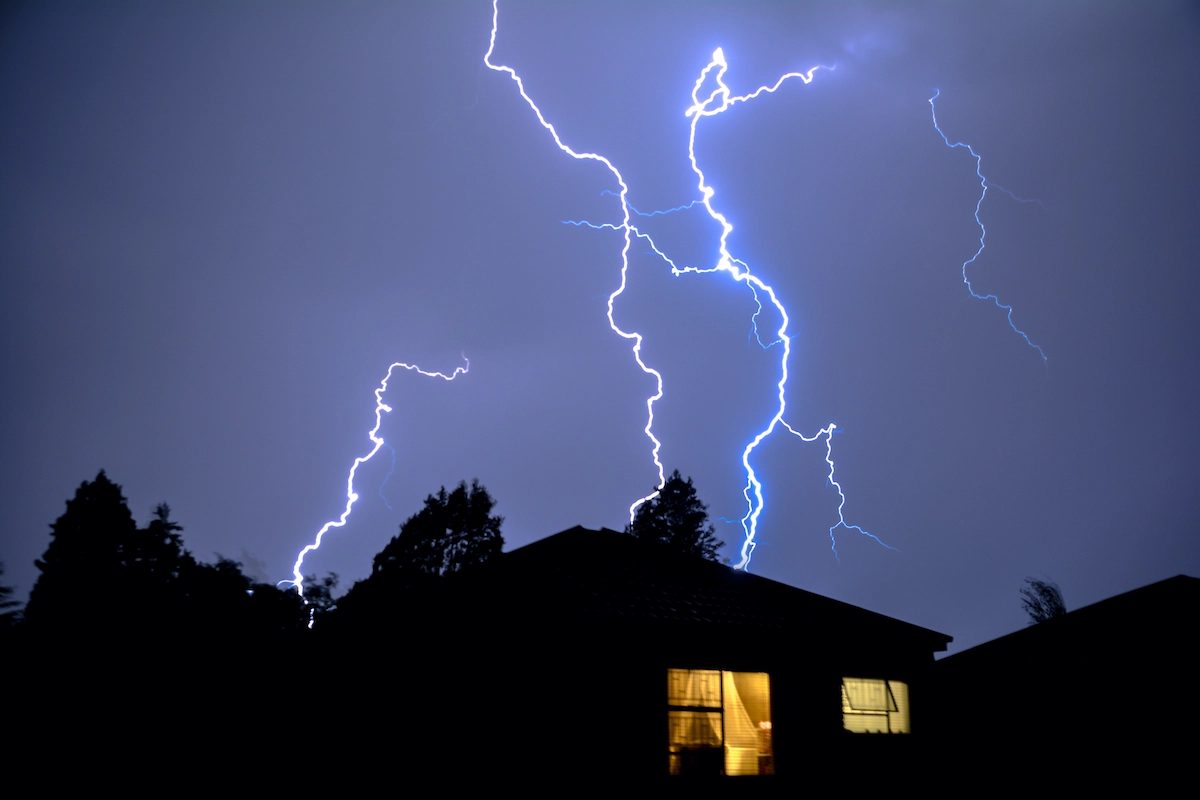 A nighttime scene shows multiple bright lightning bolts illuminating a dark sky above a silhouette of houses. One house has a window with warm light shining through, contrasting with the stormy, electrifying sky—a view familiar to anyone dealing with Houston roofing after such dramatic weather.