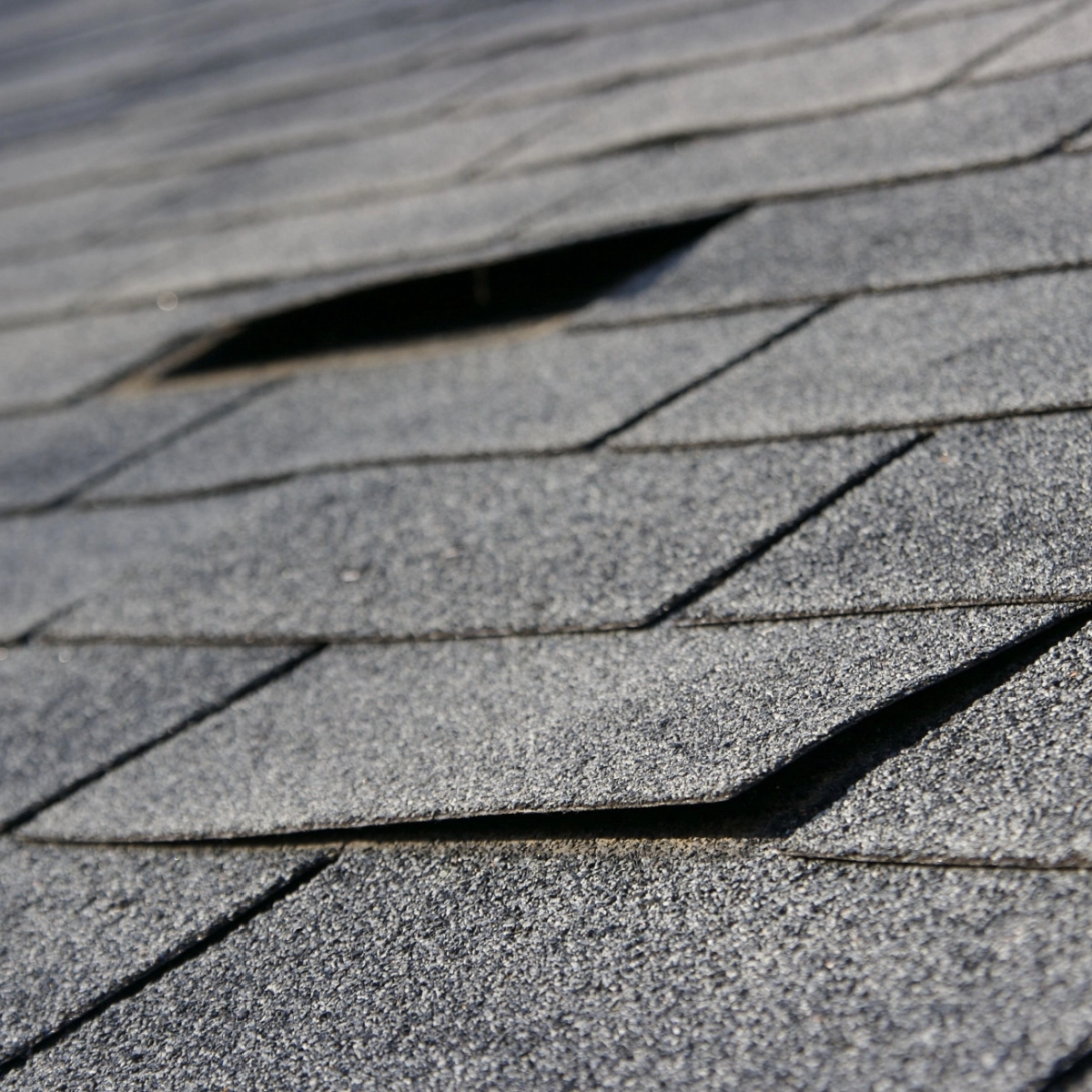 Close-up of a gray asphalt shingle roof with several shingles lifting and peeling, revealing potential damage and the need for a Houston roof replacement.