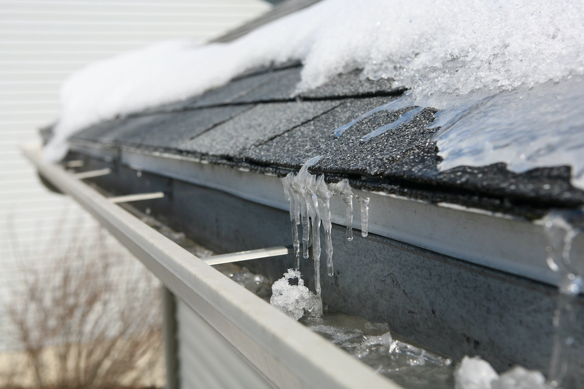Close-up of a house roof with a snow-covered shingle and gutter. Icicles hang from the edge of the roof, while the gutter is filled with ice and some debris. Houston roof experts recommend regular maintenance to prevent such icy buildup during winter months. The background shows a blurred wall and bare branches.