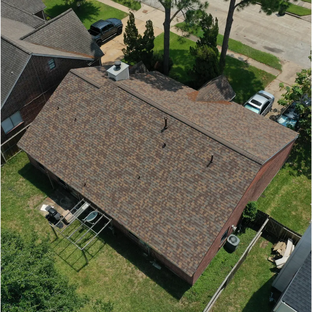 Aerial view of a brick house with a newly installed brown shingle roof. The house has an attached backyard where a partially constructed wooden structure and some garden tools are visible. The surrounding area features green lawns, trees, and parked vehicles.