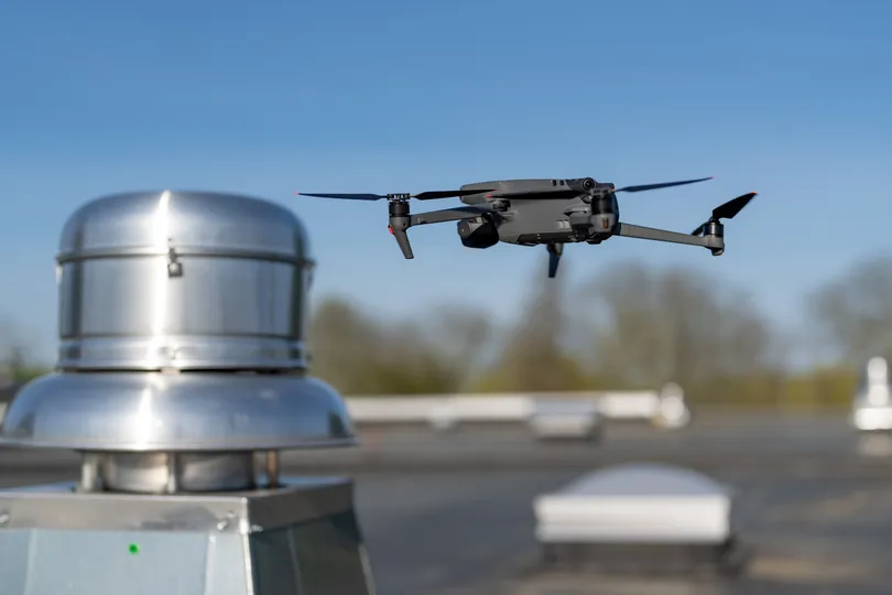 A small, black drone hovers in the air near a metallic rooftop structure with a clear blue sky in the background. The scene is set on a flat roof with blurred trees in the distance.