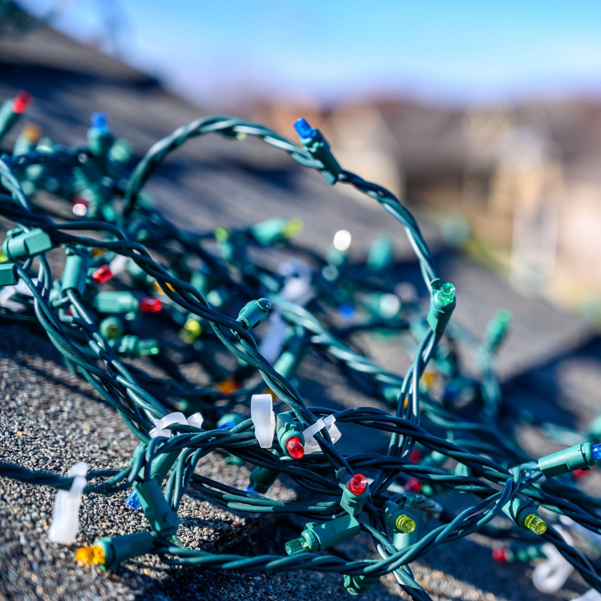 A close-up of tangled green Christmas lights with multicolored bulbs, including red, yellow, blue, and white, lying on a Sugar Land roof during an inspection. The background is blurred, showing a clear blue sky and some indistinct buildings or houses.