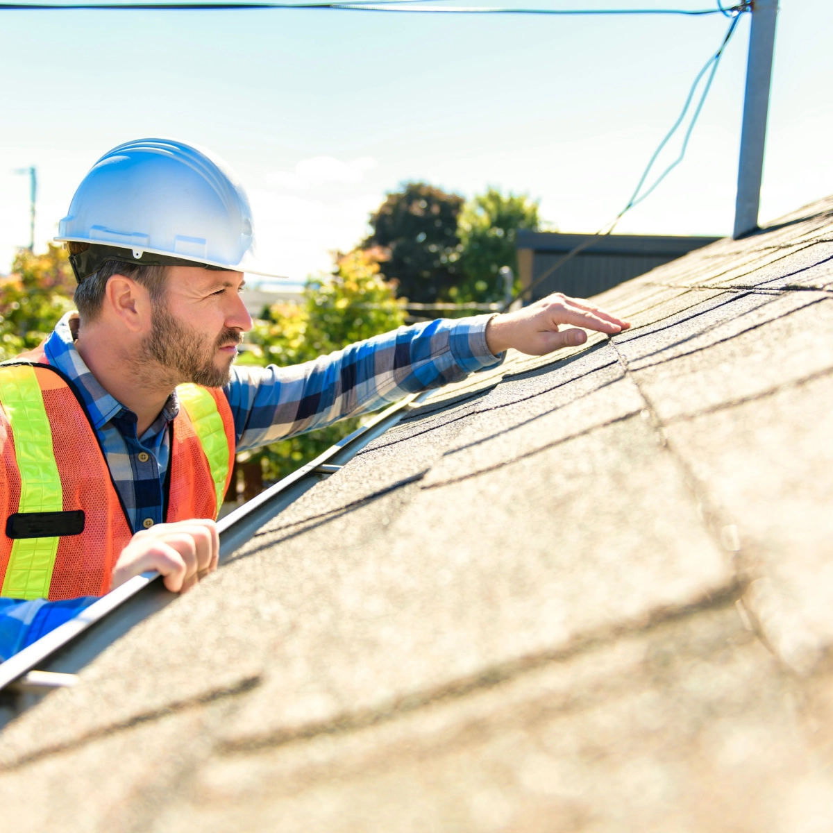 A construction worker wearing a white hard hat, orange safety vest, and plaid shirt inspects the shingles closely on a rooftop in The Woodlands on a sunny day. Trees and power lines are visible in the background.