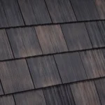 Close-up view of a wooden shingle roof. The shingles are arranged in neat, overlapping rows, with each shingle having a distinct grain and texture. The wood ranges in color from dark brown to lighter shades, creating a natural, rustic appearance.