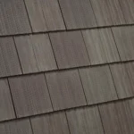 Close-up of a roof covered with brown shingles. The shingles are rectangular and arranged in an overlapping pattern, creating a uniform and textured appearance. The material has a slightly rough, grained surface, adding a natural look to the roofing.