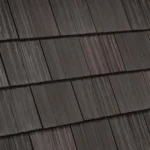 Close-up image of a roof with several dark brown shingles neatly aligned in overlapping rows, creating a textured pattern. The shingles have a slightly worn look with subtle variations in color across the surface.