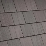 Close-up of a roof with overlapping square shingles. The shingles have a textured, ribbed pattern and are arranged in a staggered fashion, giving a neat and orderly appearance. The colors range from dark gray to brown, adding to the visual depth.
