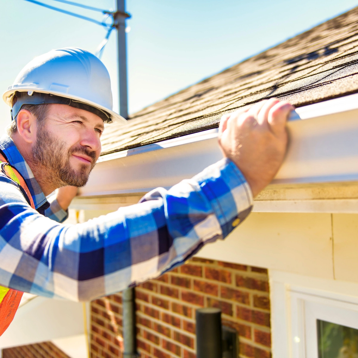 A man in a hard hat and a blue flannel shirt inspects a roof gutter, representing Rosenberg roof repair. He is standing on a ladder and looking closely at the gutter. The house has a brick exterior, and a sunny sky is in the background.