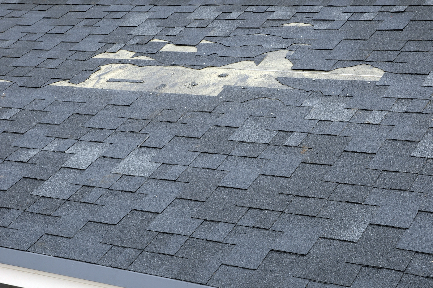A roof with gray asphalt shingles shows visible damage, likely requiring Houston roofing expertise. Some shingles are missing, exposing the underlying material. The affected area covers a significant portion in the upper middle part of the roof, indicating potential need for repair.