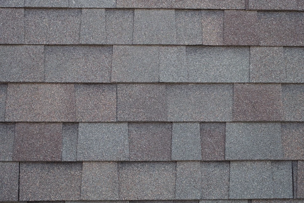 A close-up view of a roof covered with asphalt shingles highlights the meticulous work typical of a Houston roof replacement. The shingles are arranged in a staggered pattern, displaying various shades of gray and brown. The texture is rough, with a distinct granular appearance.