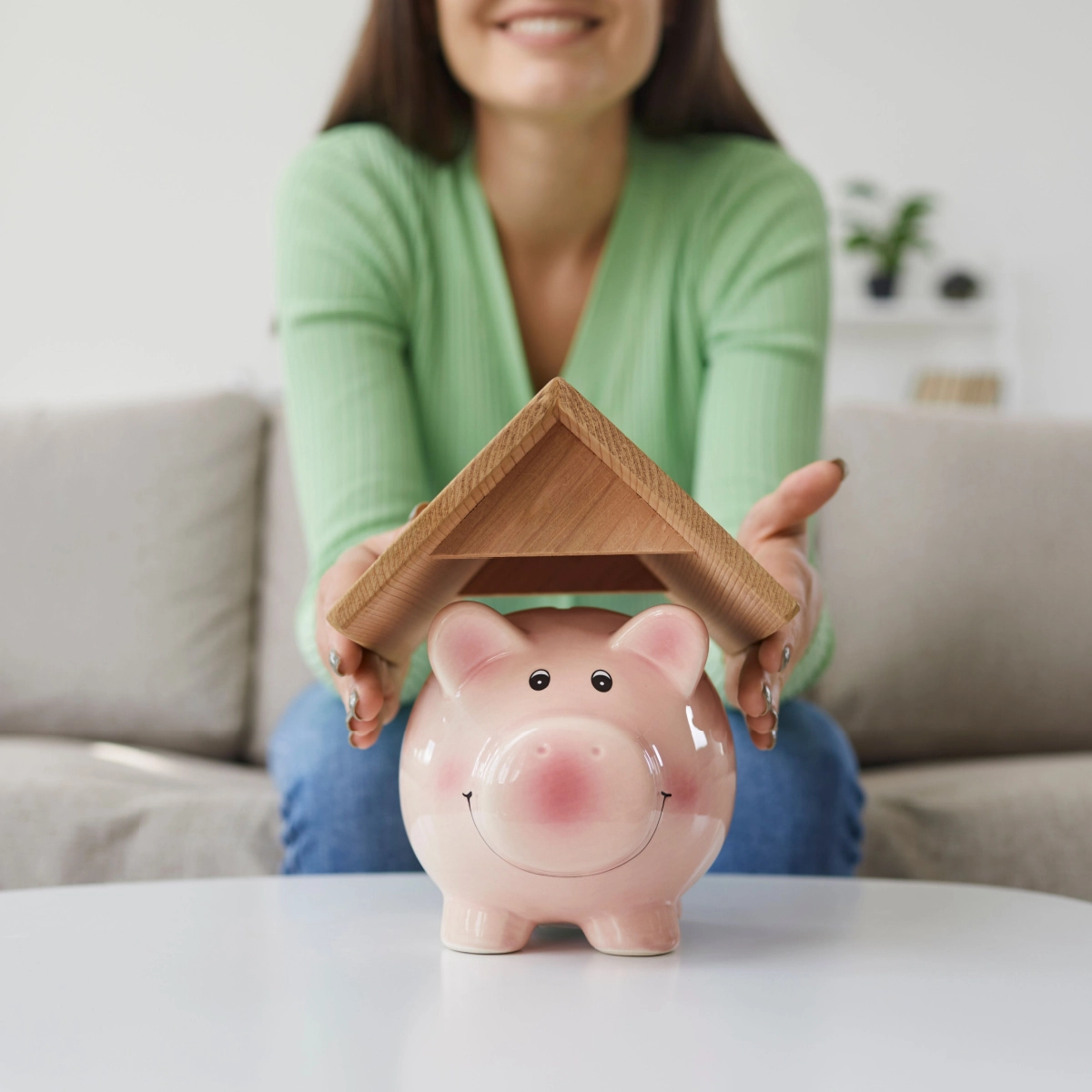 A person wearing a green sweater is sitting on a couch in the background, holding a small wooden roof above a smiling pink piggy bank placed on a white table in the foreground, symbolizing saving money or financial protection, reminiscent of Kingwood roof replacement efforts.