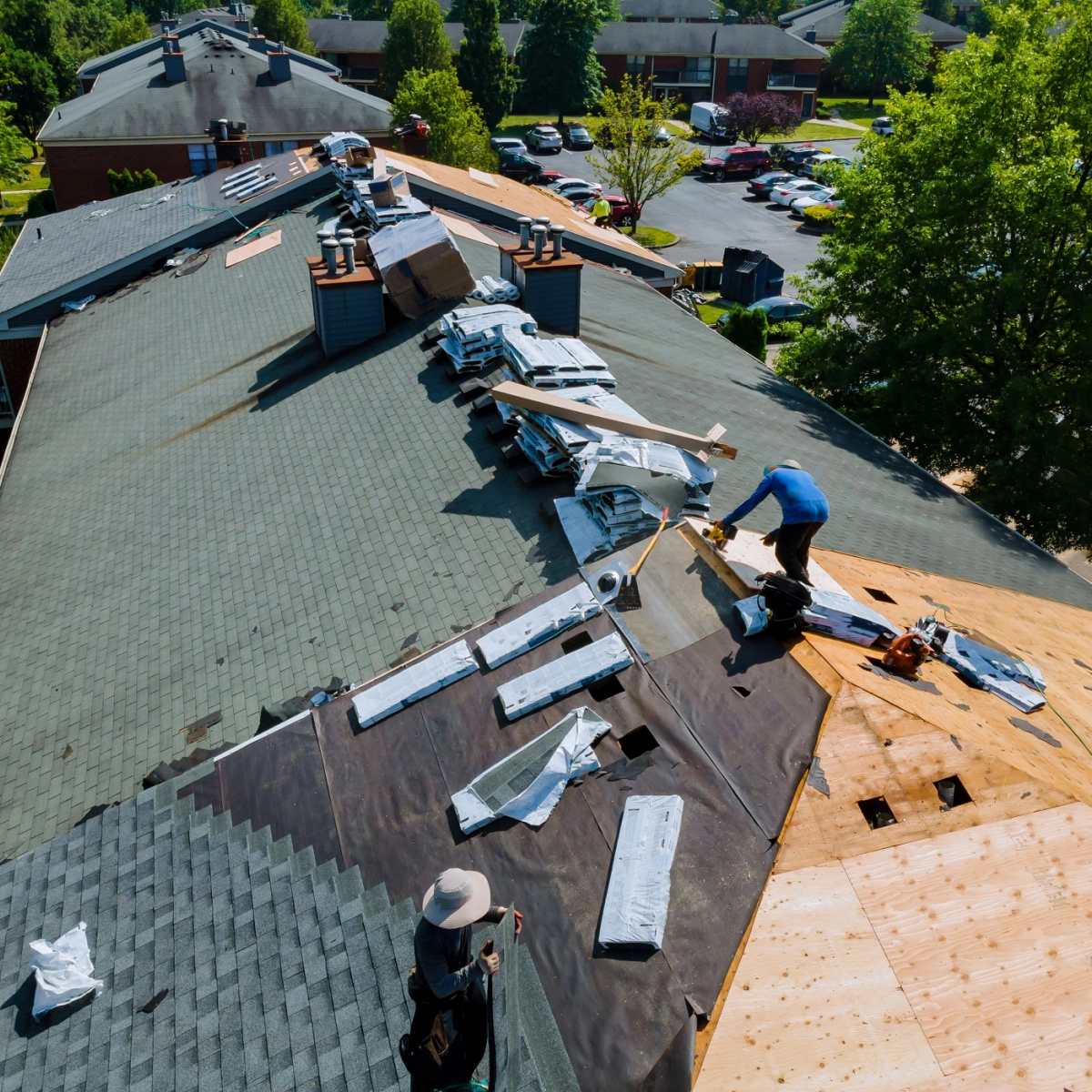 Workers repairing a roof on a large building in Tomball are laying down new shingles and tar paper. Various roofing materials are scattered around, with tools and equipment visible. Trees and parked cars are in the background on a sunny day, showcasing the thorough Tomball roof replacement process.