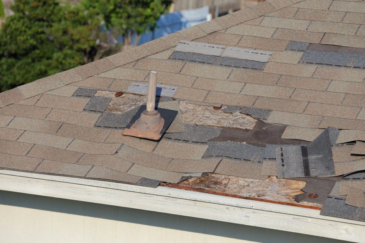 A roof with missing and damaged shingles, exposing underlayment and wood decking, highlights the need for Pearland roofing expertise. A metal vent pipe protrudes from the roof amidst various patches and mismatched shingles. Trees and blurred background structures add to the scene's urgency for repair.