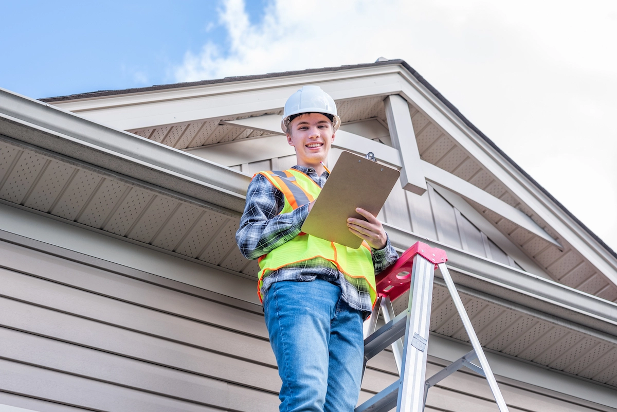 A construction worker, wearing a white hard hat, high-visibility safety vest, and plaid shirt, stands on a ladder in front of a house. He holds a clipboard and pen, smiling towards the camera with a partially cloudy sky in the background—an ideal representation of Houston roofing professionals at work.