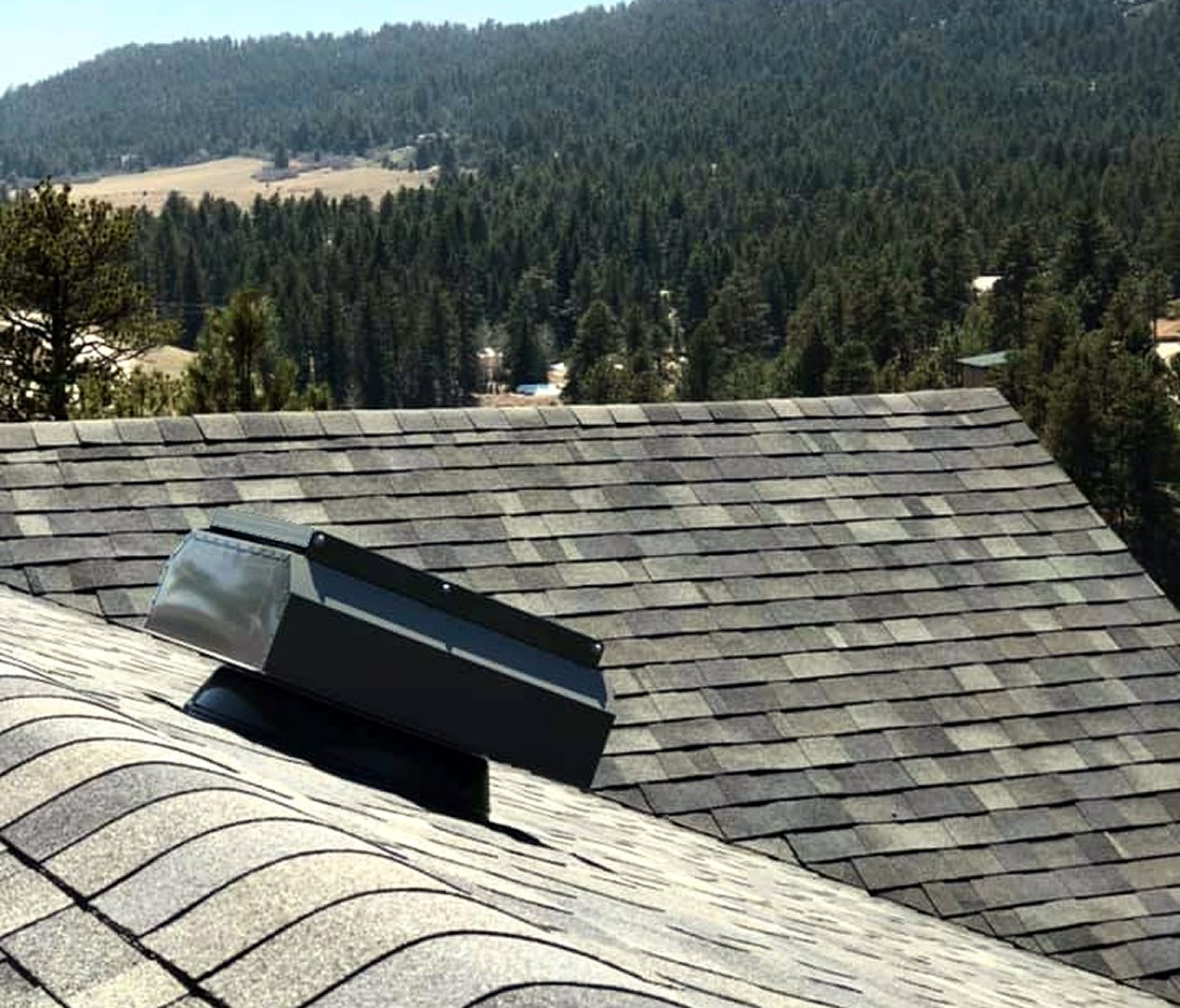 A rooftop vent on a shingled roof with a scenic background of densely forested hills and distant mountains under a clear sky. The view suggests a serene, natural setting.
