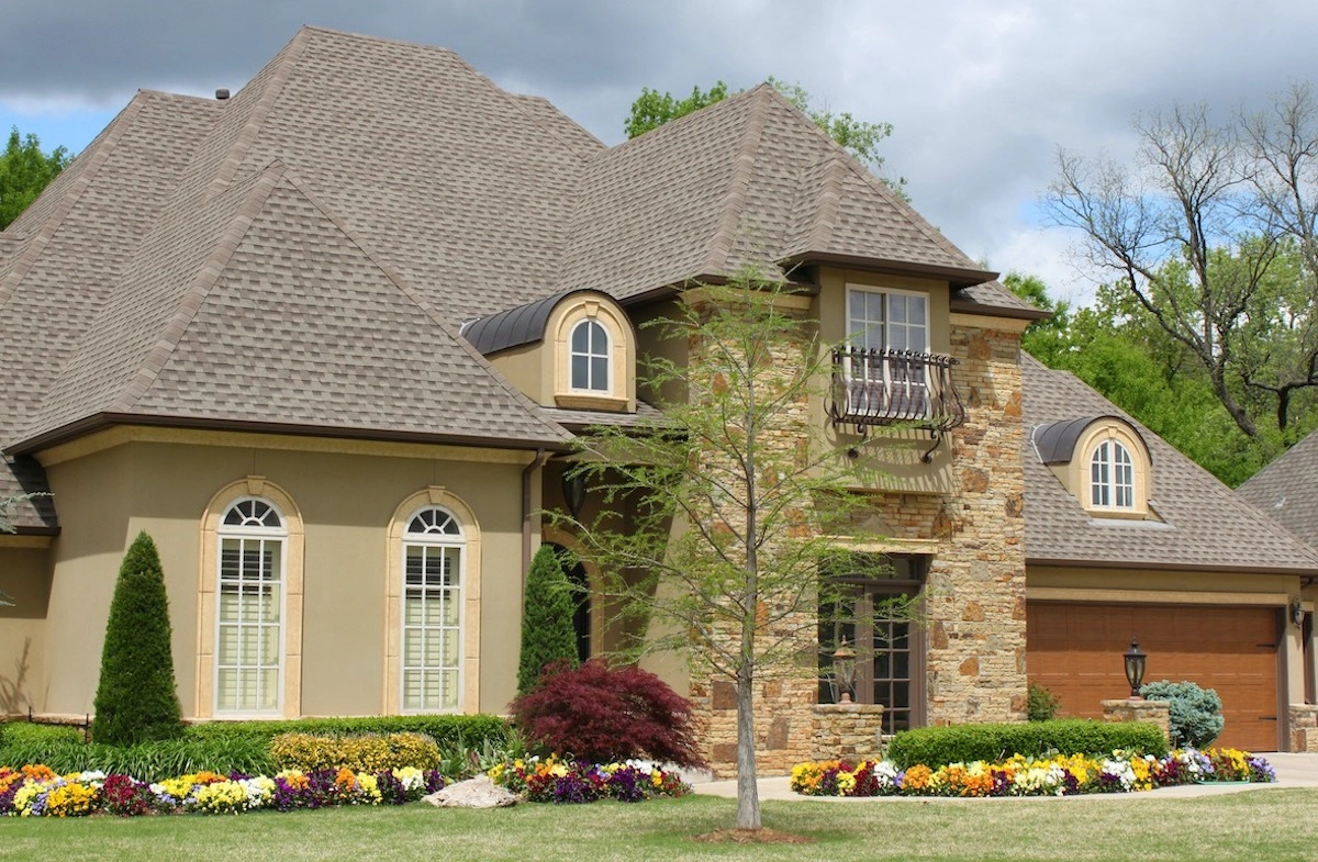 A tan, two-story house with multiple gables and a stone accent facade features large, arched windows and a small balcony. With the impeccable craftsmanship of Cinco Ranch roofing, the front yard is landscaped with multicolored flowers, shrubs, and trees. The sky is partly cloudy, and trees line the background.