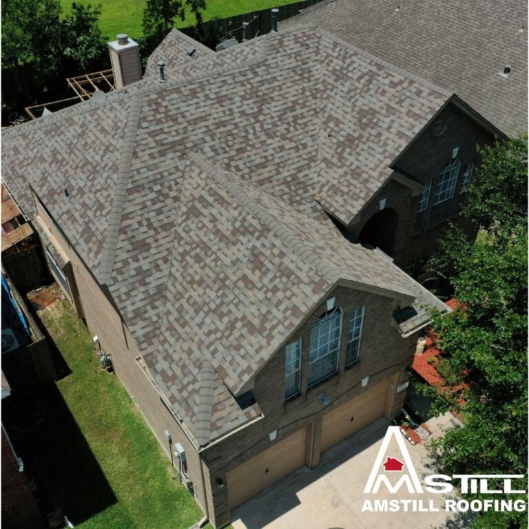 Aerial view of a residential home with a newly installed brown shingle roof. The house has a brick exterior, multiple roof peaks, and a three-car garage. Trees and a neighboring house with a similar shingle roof are visible. Average Roof Replacement Cost In Houston info in the corner, above the "Amstill Roofing" logo.