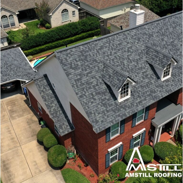 Aerial view of a two-story brick house with a newly installed grey shingle roof, featuring dormer windows. Well-maintained shrubs and a driveway are visible near the house, highlighting the home's value. "Amstill Roofing" logo is positioned in the bottom right corner of the image. Average Roof Replacement Cost In Houston.
