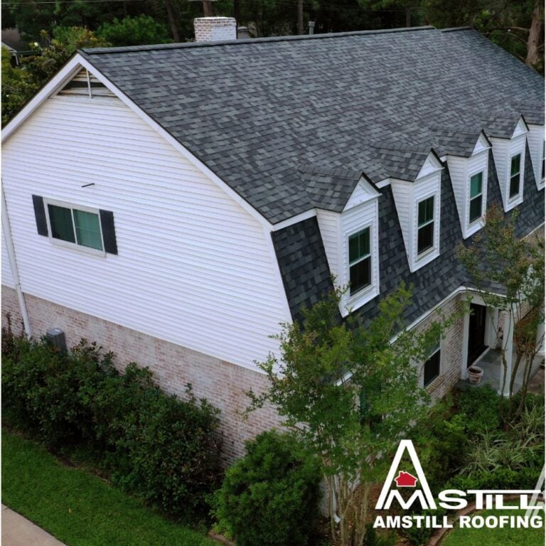 A two-story house with white siding and a checkered gray shingle roof. The house has dormer windows on the upper floor and a chimney. Trees and shrubs surround the house, exemplifying quality craftsmanship. The Amstill Roofing logo appears in the bottom right corner, highlighting the average roof replacement cost in Houston.