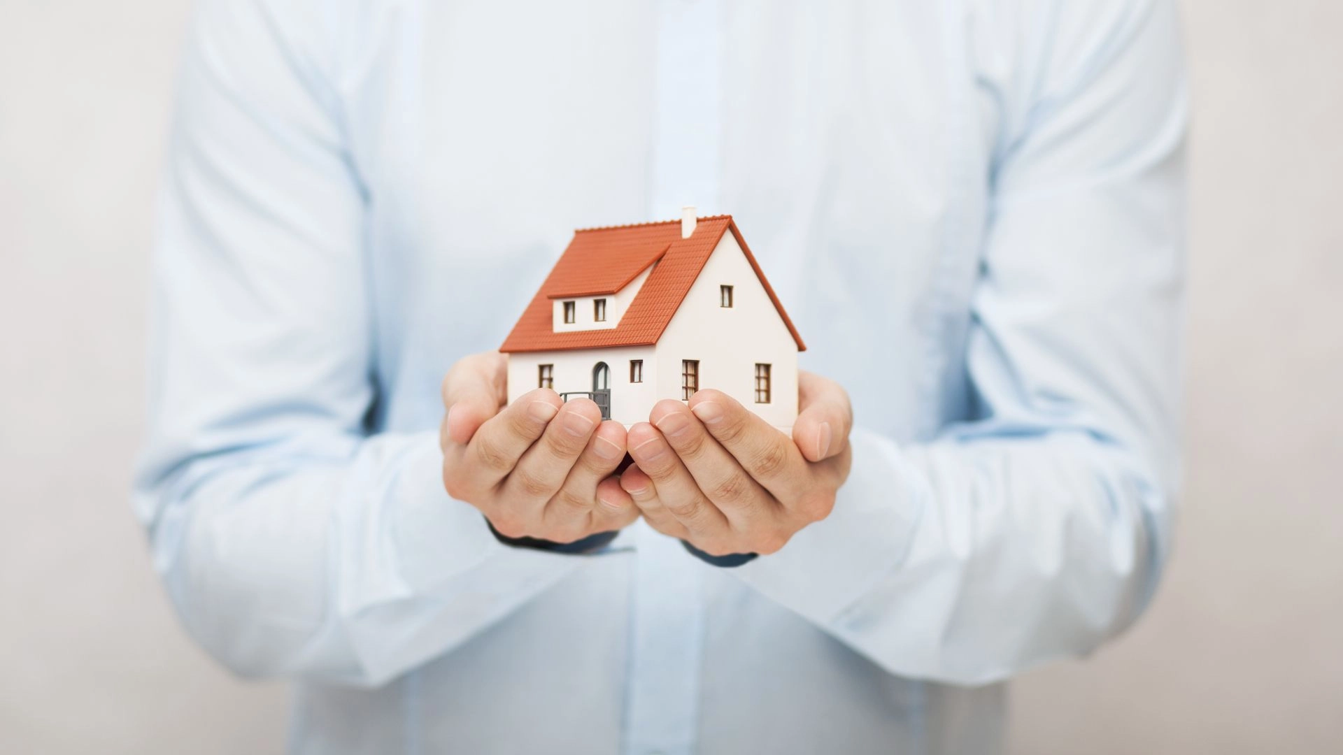 A person in a light blue shirt is holding a small model of a house with red roof and white walls. The person's face is not visible. The background is plain and light-colored.