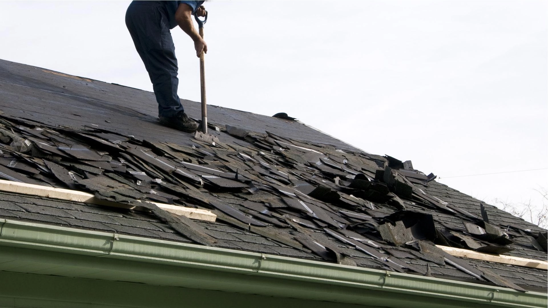 A person in dark clothing is standing on a roof in Houston, using a tool to remove old, damaged shingles. The roof is covered with partially removed shingles and debris. The person is working at an angle on a steep section of the Houston roofing project.