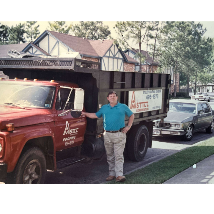 A man in a blue shirt stands smiling next to a red truck with "Still Roofing" written on the door. The truck is parked on a residential street, and there are houses with trees and parked cars in the background. The man has his hand resting on the truck's door.